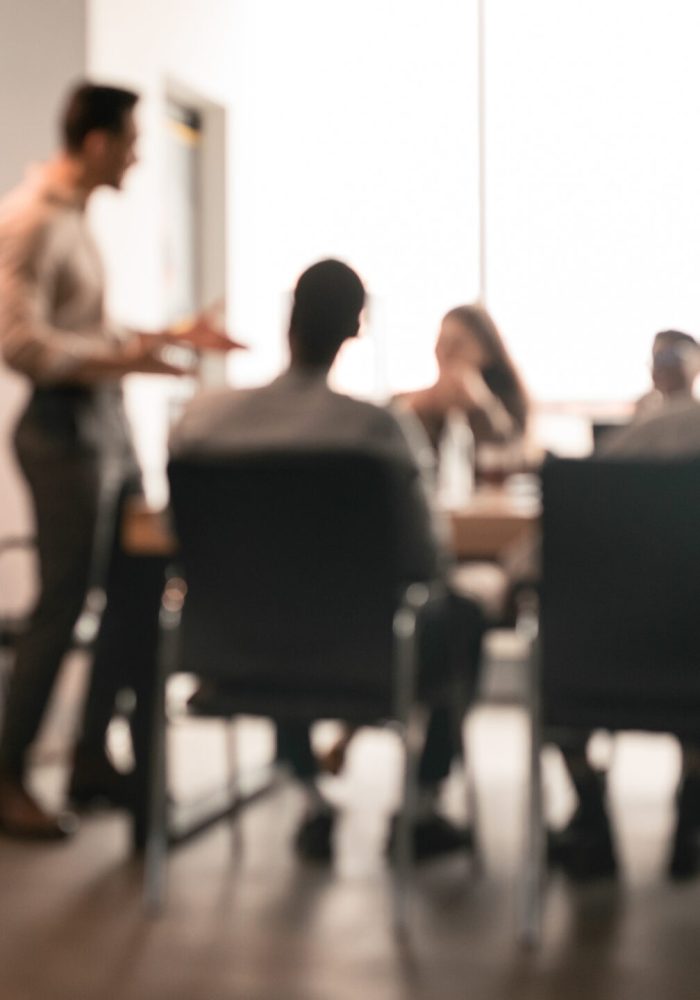 Colleagues having meeting in boardroom, businessman giving speech, blurred photo Business Presentation, Blurred Background. Businessman Giving Speech During Seminar With Coworkers In Office, Standing At Desk In Boardroom, Diverse People Sitting At Table And Listening To Speaker